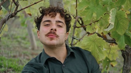 Portrait of young man winegrower sitting alone under vine bush eating grapes. Italian professional viticulturist relaxing after work day on plantation close up. Vineyard worker tasting sweet berries.