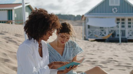 Photo for Two girls reading book on summer beach picnic. Happy lesbian couple talking enjoying novel in sunlight. Carefree multiethnic friends discussing literature on sandy coast. Vacation weekend concept. - Royalty Free Image