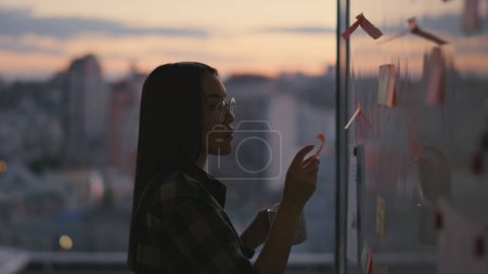 Photo for Woman silhouette reading stickers at sunset office close up. Unknown hipster lady pondering project ideas at evening place alone. Attentive girl applying notes papers at panoramic windows apartment - Royalty Free Image