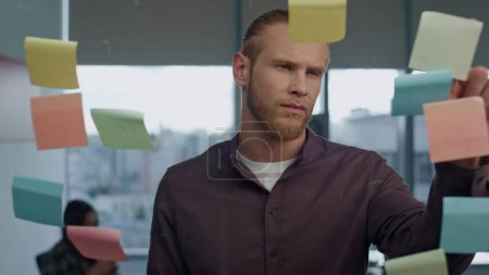 Photo for Thoughtful man pondering sticker wall office closeup. Young freelancer preparing tasks on sticky notes transparent board. Serious startuper brainstorming creative ideas at big windows coworking - Royalty Free Image