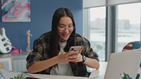 Photo for Cheerful woman messaging smartphone at office closeup. Smiling girl sitting desk answering email at blue wall room alone. Z-generation student browsing social media at panoramic windows workplace - Royalty Free Image