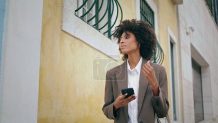 Téléchargez les photos : Relaxed carefree girl walking city street holding smartphone. Happy young african american woman using mobile phone taking picture building door. Attractive tourist lady exploring town architecture. - en image libre de droit