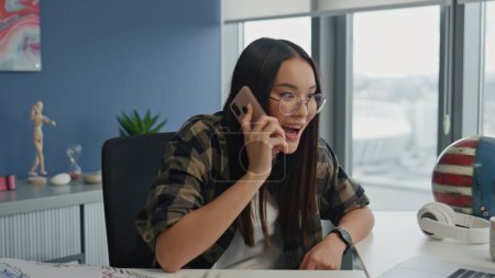 Photo for Excited manager gossiping phone in office interior closeup. Smiling woman calling mobile at hipster remote workplace. Friendly specialist holding cellphone talking emotionally at city view apartment - Royalty Free Image