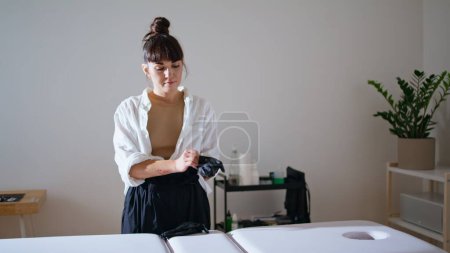 Photo for Tattoo girl wearing rubber gloves at workplace. Focused tattooist preparing receiving client at studio. Serious woman standing at couch place pulls on protective equipment. Creative occupation concept - Royalty Free Image