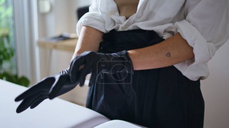 Photo for Woman hands wearing gloves at salon close up. Unrecognizable tattooist preparing to client session at remote workplace. Tattoo artist fingers pulls on protective rubber equipment at studio alone - Royalty Free Image