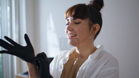 Photo for Smiling tattooist wearing rubber gloves at studio closeup. Positive tattoo girl preparing receiving client at workplace. Cheerful woman pulls on protective equipment. Creative occupation concept - Royalty Free Image