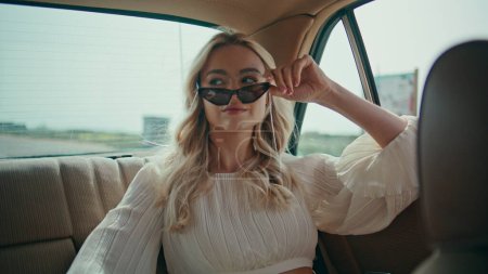 Photo for Girl travelling at car backseat looking window on road view close up. Carefree attractive woman passenger riding in retro automobile in white dress. Blonde sitting auto back seat taking off sunglasses - Royalty Free Image