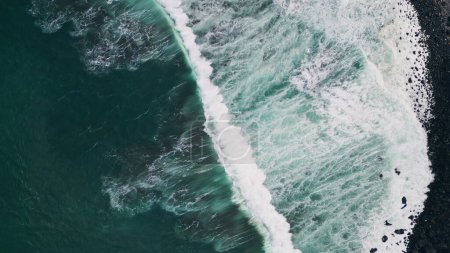 Téléchargez les photos : Mousse océanique éclaboussant sur la plage sombre et pierreuse vue sur drone roulant. Ondes agitées qui lavent la mousse du bord de la mer au ralenti. Belle eau de mer frappant la côte volcanique noire. Paysages marins nature - en image libre de droit