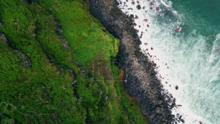 Téléchargez les photos : Océan surf éclaboussures sur les falaises moussues nature drone vue de dessus. Ondes moussantes orageuses lavant plage pierreuse sombre s'écrasant rivage volcanique au ralenti super. Belle eau de mer frappant la côte marine noire - en image libre de droit