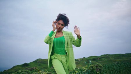 Mujer afroamericana bailarina moderna moviendo cuerpo sensualmente en el prado nublado. Chica rizada inspirada realizando danza contemporánea de pie en hierba verde. Expresiva jovencita bailando suavemente al aire libre