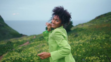 Smiling playful african american girl moving body energetically on cloudy field closeup. Happy cheerful woman dancer performing modern choreography on green flowers meadow. Lady dancing in slow motion