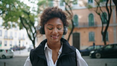 Relaxed african american woman enjoy song in wireless headphones standing city street close up. Happy smiling girl listening music in modern earphones outdoors. Pretty lady moving head rhythmically.