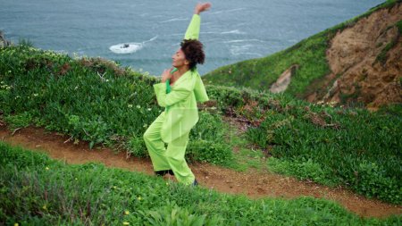 African american young woman choreography dancing at evening coast. Professional dancer performing modern dance style on gloomy nature seashore. Curly lady in green suit moving gracefully outdoors.