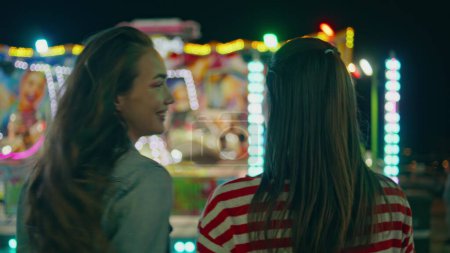Funfair girls watching illuminated carousel rear view. Happy teenage friends hang out enjoying summer evening at amusement luna park. Two smiling cheerful teenagers having fun resting city festival.