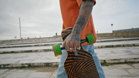 Unknown tattooed man skater walking with modern longboard on street. Young longboarder carrying skate board in hand going at urban backdrop. Unrecognizable skateboarder strolling outdoors cloudy day.