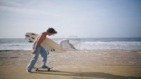 Sporty cool man riding skateboard with surfboard at sunny quay road. Attractive half naked skateboarder accelerating on boardwalk sea beach holding surf board. Tattooed guy enjoy hobby in slow motion.