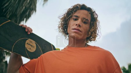 Photo for Cool skateboarder holding skate board portrait in cloudy tropical summer view. Attractive curly guy skater with longboard looking camera outdoors close up. Trendy hipster enjoy active hobby on street - Royalty Free Image