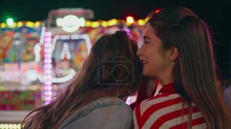 Closeup happy girls sharing secrets at funfair. Two friends gossiping laughing at blinking colorful carousel. Cheerful teenagers talking having fun at luna amusement park. Friendship bonding concept