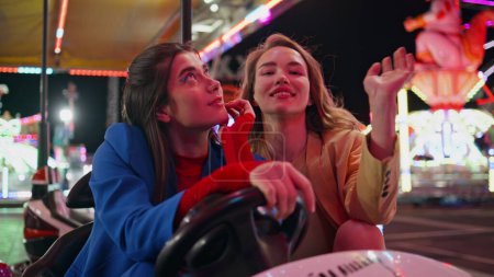 Girls rest amusement park in illuminated carousel closeup. Two smiling friends enjoying summer evening bumper car ride at funfair. Happy joyful models girlfriends hang out together in neon lights.