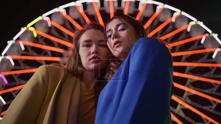 Two girls posing ferris wheel at night closeup. Gorgeous friends at illuminated neon carousel in amusement luna park. Serious urban models rest funfair together. Attractive female couple at dark sky