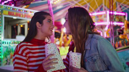 Happy teenagers sharing secrets at illuminated carousel. Cheerful friends gossip hang out together in amusement luna park. Excited smiling girls talk having fun on weekend funfair. Female friendship