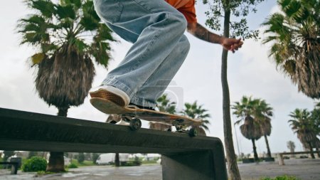 Inconnu skateboarder actif pratiquant kickflip de banc de rue gros plan. Jeune homme jambes sautant à longboard entraînement tour au ralenti. Patineuse sportive pratiquant un passe-temps extrême près des palmiers tropicaux