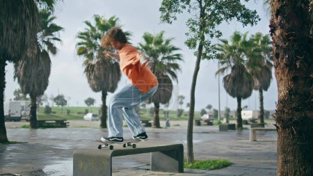 Patineur cool pratiquant le tour extrême de skateboard à la rue de la ville. Jeune homme sautant du banc avec skateboard dans un parc tropical. Hipster sportif effectuant kickflip à l'extérieur au ralenti. Loisirs actifs.