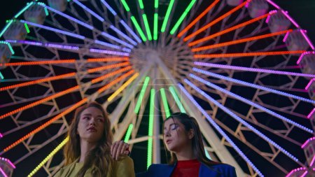 Closeup beautiful girls posing at colorful ferris wheel. Fashion models at night resting amusement luna park carousel. Two gorgeous female girlfriends lean each other looking camera at blinking lights