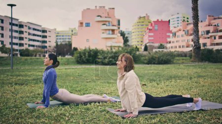 Les femmes sportives détendues pratiquant le cobra posent parc de la ville. Yogini heureux se pencher en arrière position de pose de yoga sur la surface herbeuse. Athlète femmes étirant le corps le matin. Filles flexibles pratiquant des exercices 