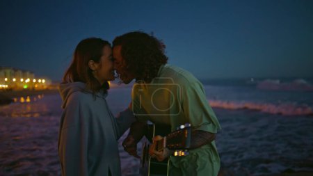Photo for Enamoured man playing acoustic guitar at night beach closeup. Curly young guy serenading at musical instrument kissing girlfriend at evening. Relaxed carefree sweethearts spending free time in nature - Royalty Free Image