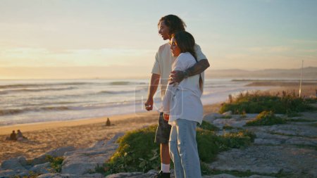 Photo for Teen couple enjoy sundown ocean view. Happy emotional youngsters communicating on sea beach nature. Young lovers hugging on summer trip looking marine horizon. Beautiful romantic vacation together - Royalty Free Image