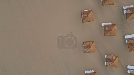 Panoramic aerial view empty sun loungers and straw parasols on seashore. No people beach early morning zoom out. Top shot recliners and umbrellas at sandy surface. Summer coastline vacation concept