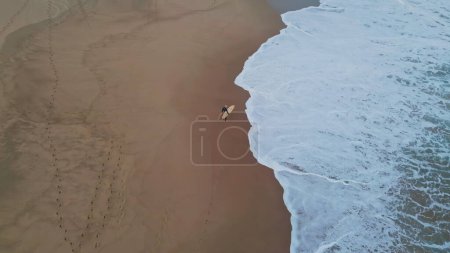 Drone shot unknown surfer walking in sandy beach waiting waves. Wonderful marine view surfboarder carrying board stepping wet sand with foamy ocean water slow motion. Active hobby leisure concept 