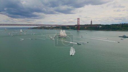 Lisboa cityscape floating yachts at cloudy day drone shot. Many boats sailing in calm still water passing green island. Puffy dark clouds gathering over city area before rain. Famous 25th April bridge
