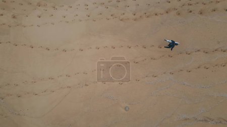 Drone shot surfboarder walking sand holding professional surfboard. Unrecognizable person strolling seashore leaving footprints top view. Unknown surfer relaxing after active summer hobby alone.