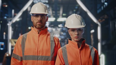 Two professional engineers posing at modern production workshop in uniform close up. Serious couple factory workers standing in large plant warehouse. Technical specialists looking camera confidently.