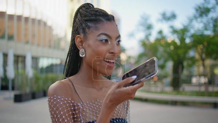 Femme élégante enregistrant un message vocal sur le microphone du smartphone à proximité de la rue. Heureuse femme afro-américaine parlant dans un haut-parleur de téléphone à l'aide de l'application mobile assistant. Concept de technologie moderne 