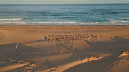Panoramic view tropical beach ocean surf. Aerial empty recliners and umbrellas on seashore morning zoom on. Drone shot sun loungers and parasols at sandy surface. Summer coastline vacation concept