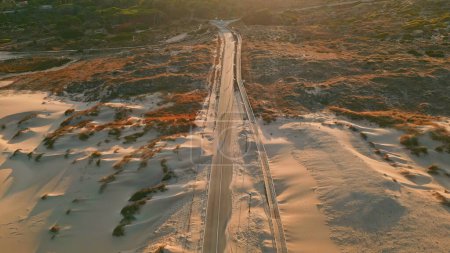 Golden sunlight sandy trail drone view. Aerial beautiful coast road between dunes. Green picturesque hills with sand. Panoramic evening summer scenic empty highway without cars. Journey and nature