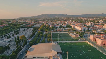 Drone view peaceful small town with beautiful green football stadium. Urban well-groomed districts under evening sunlight. Aerial shot cozy city neighborhood with grass soccer field calm summer day.
