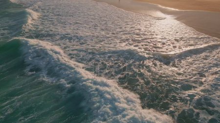 Téléchargez les photos : Sable de lavage de l'océan mousseux à proximité du soleil doré. Surf de mer orageux couvrant la plage de sable au ralenti. Vue cinématique pulvérisation blanche moussant dans les eaux peu profondes roulant sur le littoral. Nature concept de fond - en image libre de droit