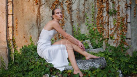 Herrliches Modell posiert beim Herbstlaub auf alten Wandzoom. Elegante attraktive Frau, die auf verlassenen Steinen sitzt und verträumt aussieht. Stilvolles Mädchen im weißen Kleid fühlt Einsamkeit am Abend in der Natur