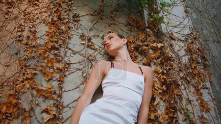 Frau stützt sich auf altes, mit gelbem Herbstlaub bedecktes Gebäude. Posh schöne Modell sieht Kamera selbstbewusst in weißen eleganten Kleid. Attraktive luxuriöse Mädchen posieren auf der Natur. Modekonzept.