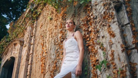 Entspannt lehnt eine Frau an einem mittelalterlichen Gebäude, das Herbstlaub bedeckt. Selbstbewusst posiert das schöne Model im weißen Kleid vor einem verlassenen Palast. Wunderschönes Mädchen, das vor die Kamera blickt und Sinnlichkeit ausdrückt.