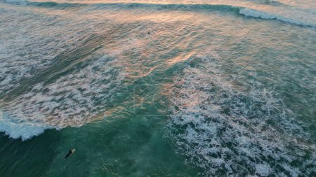 Drone view foamy sea water swinging unknown surfers under evening summer sunlight. Extreme surfboarders waiting high waves at vacation. Beautiful ocean swell rolling to shoreline in super slow motion.