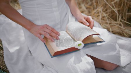 Lady fingertips touching book pages in rye field close up. Unrecognizable white dress woman sitting blanket reading literature at fresh air summer picnic. Unknown girl holding novel with tender flower