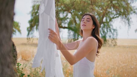 Sensual lady hanging laundry at meadow closeup. Romantic brunette woman putting clean white linen on rope enjoying washing day in rye field. Portrait girl looking at camera posing at countryside