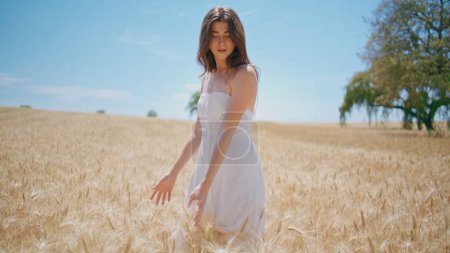 Relaxed woman strolling sunlight farmland. Smiling lady enjoying nature landscape walking morning meadow. Serene beautiful model hand touching wheat stalks at agriculture harvest. Simple pleasures 
