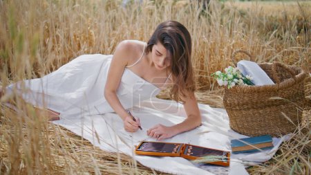 Dreamy girl sketching album relaxing blanket at rye landscape. Attractive model laying dry spikelets nature drawing alone at morning time. Thoughtful woman enjoying creative hobby at fresh air 