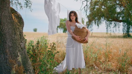 Mujer joven sosteniendo cesta de lavandería caminando jardín de hierba seca. Chica sensual calma mirando cámara paso lentamente en el campo de espiguillas de trigo. ama de casa rural llevando alforja de mimbre cruzando pradera de centeno